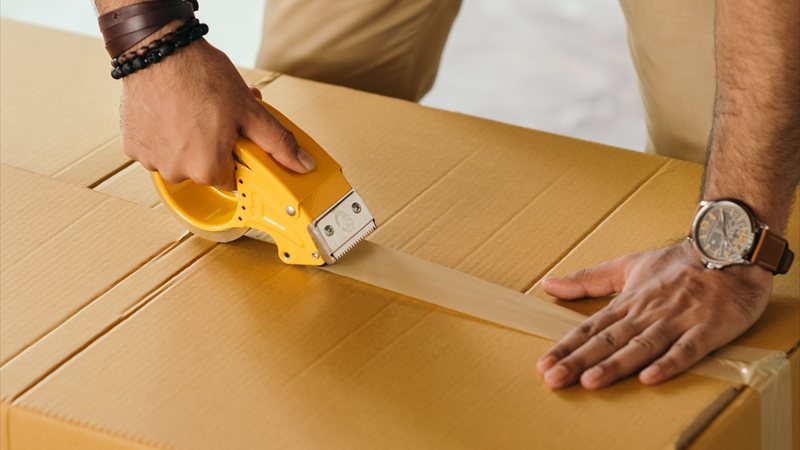 Mann verschließt einen Umzugskarton sorgfältig mit einem Klebebandabroller